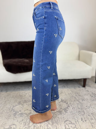 A person wearing blue denim jeans with small white embroidered floral details, shown from the waist down in a living room setting with a white sofa and patterned rug.