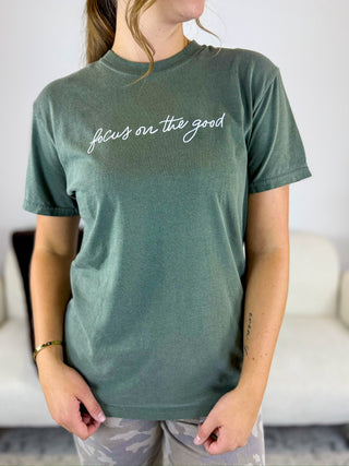 A woman wearing a sage green short-sleeve t-shirt with the phrase 'focus on the good' printed in white script across the chest, paired with camouflage leggings.