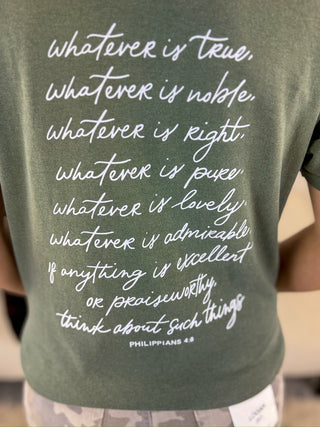 A close-up of a person wearing an olive green t-shirt with a white printed inspirational Bible verse from Philippians 4:8 on the back.