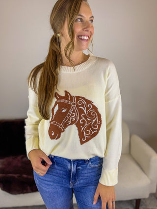 A woman wearing a cream-colored knit sweater with a brown horse head graphic on the front, paired with blue jeans, standing in a living room setting.
