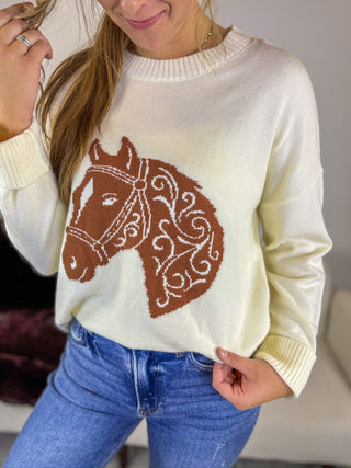 A woman wearing a cream-colored knit sweater with a brown horse head graphic on the front, paired with blue jeans.