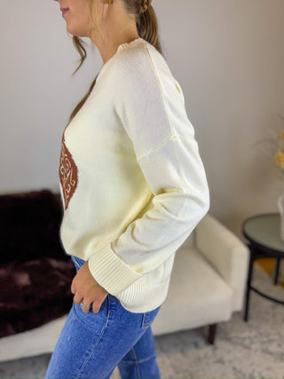A woman wearing a cream-colored knit sweater with a brown embroidered circular design on the front, paired with blue jeans, standing in a living room setting.