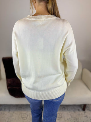 A woman viewed from behind wearing a cream-colored knit sweater and blue jeans in a living room setting.