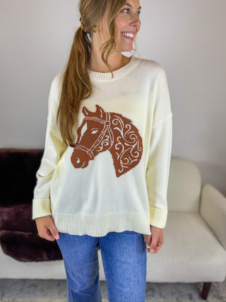 A woman wearing a cream-colored knit sweater with a brown horse head graphic on the front, paired with blue jeans, standing in a living room setting.