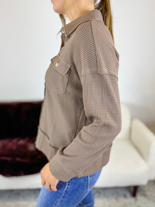 A woman wearing a brown waffle-knit button-up jacket with chest pockets, paired with blue jeans, shown in a side profile view in an indoor setting.