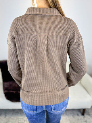 A person shown from the back wearing a taupe waffle-knit cropped jacket with pleated back detail and blue jeans, standing indoors.