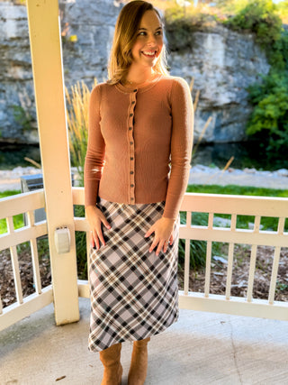 A woman stands on a porch wearing a ribbed long-sleeve button-front cardigan in a warm camel brown, paired with a plaid midi skirt in beige, gray, and brown tones.