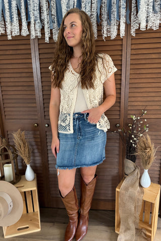 Woman wearing a lace top, denim skirt, and brown knee-high boots standing in front of wooden shutters.