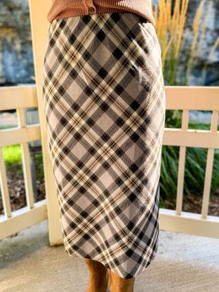 A plaid midi skirt in beige, black, and white, worn by a model standing on a porch with a white railing and plants in the background.