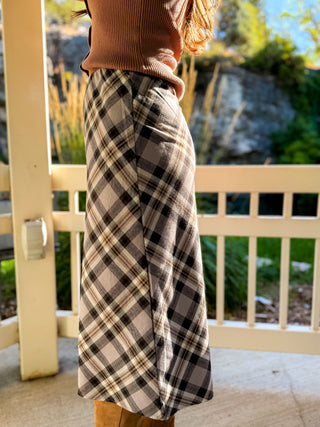 A person wearing a wide-leg plaid skirt in beige, brown, and black, paired with a brown ribbed top and tan boots, standing on a porch with a garden background.