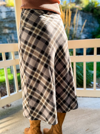 A person wearing a knee-length plaid skirt in beige, black, and gray tones, paired with tan boots, standing on a porch with a white railing and outdoor greenery in the background.