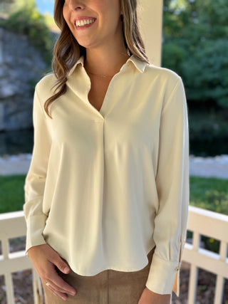 A woman wearing a cream-colored long-sleeve button-down blouse, standing outdoors on a porch with a white railing and green foliage in the background.