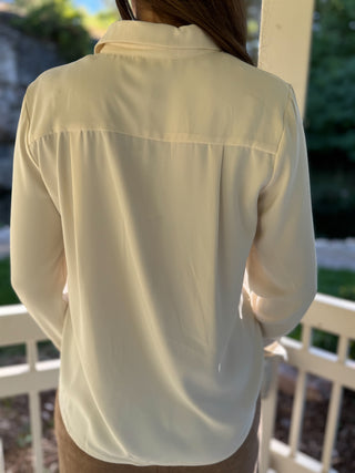 A woman viewed from behind wearing a light cream or off-white long-sleeve button-up shirt, standing outdoors on a porch with a white railing and greenery in the background.
