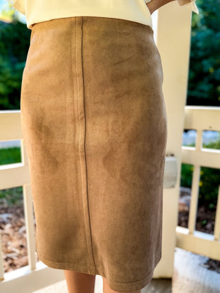 A tan faux-suede pencil skirt worn by a model standing on a white porch with green foliage in the background.