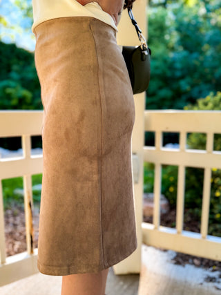 A person wearing a knee-length tan suede or faux-suede skirt, standing outdoors on a porch with a white railing and green foliage in the background.