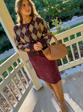 A woman stands on a porch wearing a burgundy argyle cardigan, a burgundy midi skirt, tan suede boots, and carrying a tan leather handbag.