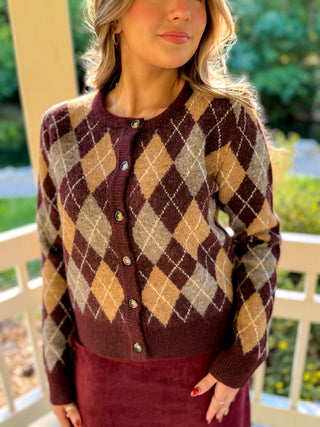 A woman wearing a cropped button-front cardigan with an argyle pattern in dark burgundy, tan, and beige, paired with a burgundy skirt, standing on a porch with greenery in the background.