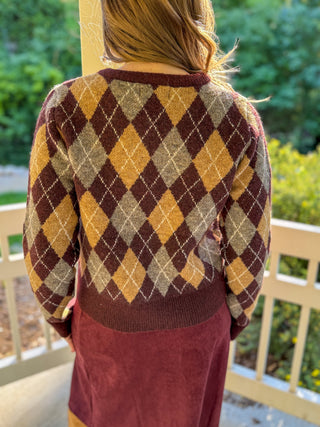 A person viewed from behind wearing a vintage-style argyle sweater with diamond patterns in mustard yellow, burgundy, and beige, paired with a matching burgundy skirt, set against an outdoor porch background.