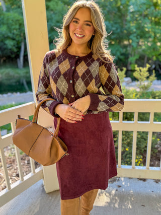 A woman stands on a porch wearing a burgundy and tan argyle cardigan over a burgundy midi dress, paired with tan boots and holding a tan handbag.