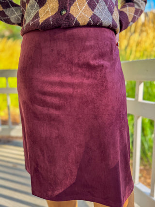 A person wearing a knee-length burgundy suede skirt and a patterned knit top, standing outdoors on a porch with a white railing and blurred green and yellow foliage in the background.
