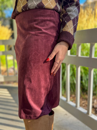 A person wearing a knee-length burgundy corduroy skirt, styled with a brown argyle sweater and tan boots, standing outdoors on a porch with a white railing.