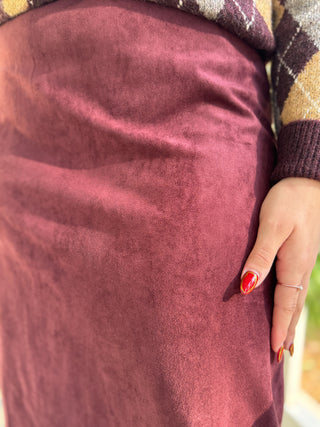 A close-up of a person wearing a dusty rose faux suede skirt, paired with a brown and beige plaid sweater. The person's hand with red nail polish rests on the skirt.