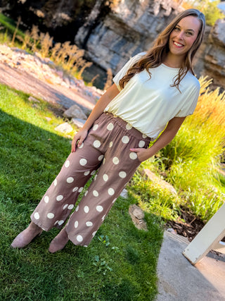 A woman posing outdoors in brown wide-leg pants with white polka dots and a cream t-shirt, set against a natural background of grass and rocks.