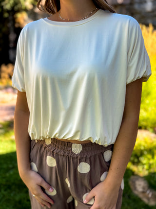 A woman wearing a cream-colored short-sleeve crewneck top and brown polka-dot pants, standing outdoors in a garden setting.