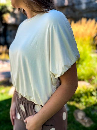 A woman wearing a loose, short-sleeve cream-colored top and brown polka-dot pants, photographed outdoors in a garden setting.