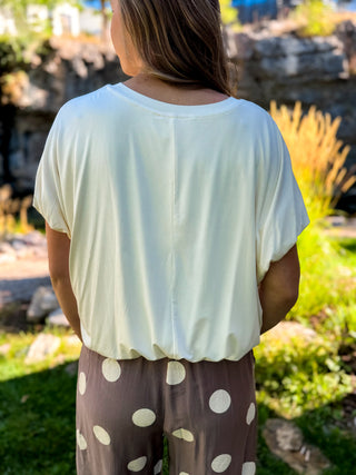 A woman viewed from behind wearing a cream-colored short-sleeve top and brown pants with white polka dots, standing outdoors in a garden setting.