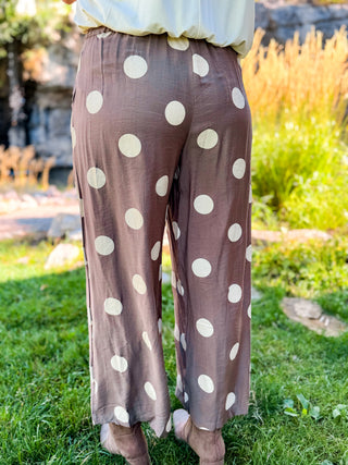A person standing outdoors on grass wearing loose, wide-leg pants in a dusty mauve-brown color with large cream polka dots. The photo is cropped from the waist down, showing the lower half of the body and the pants in a garden setting.