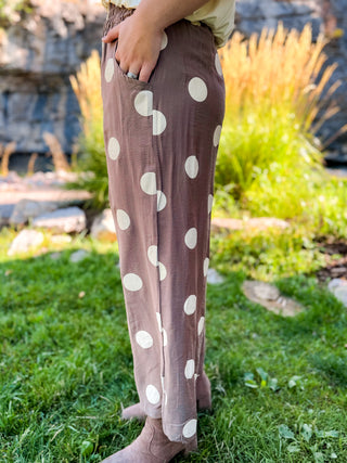 A person outdoors wearing loose, wide-leg taupe pants with large white polka dots, standing on grass with rocks and plants in the background.