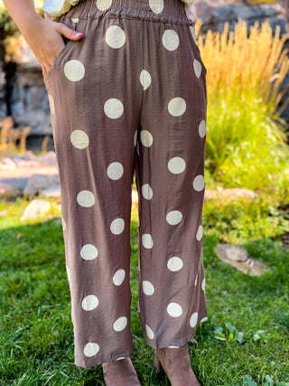 A person wearing loose brown wide-leg pants with large cream polka dots, standing outdoors on grass with a garden background.