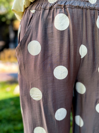 Close-up of a person wearing loose, wide-leg pants in a muted taupe-brown fabric with large white polka dots, photographed outdoors with a blurred green background.