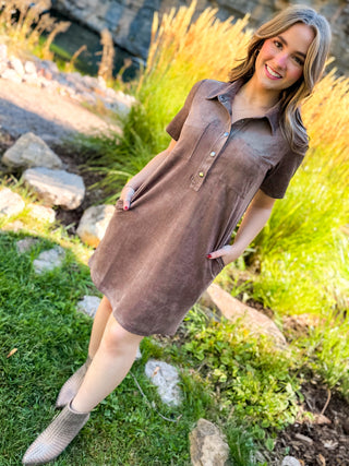 A woman wearing a brown short-sleeve button-front dress with side pockets, standing outdoors on grass with rocks and tall grass in the background.