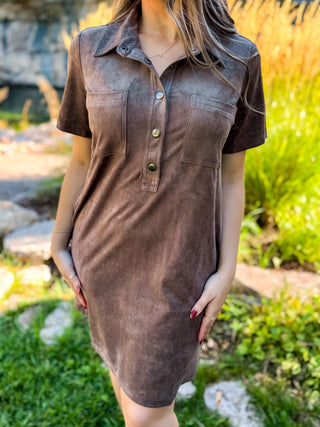 A woman wearing a brown short-sleeve collared shirt dress with a button placket and chest pockets, standing outdoors in a garden setting.