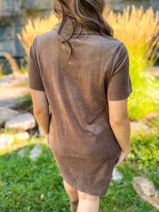 A woman viewed from behind wearing a short-sleeve brown linen dress in an outdoor garden setting.
