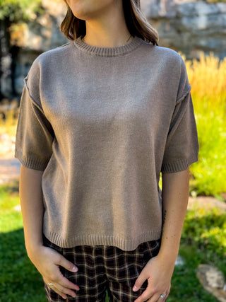 A woman wearing a taupe short-sleeve knit sweater and dark plaid pants, standing outdoors in a garden setting.