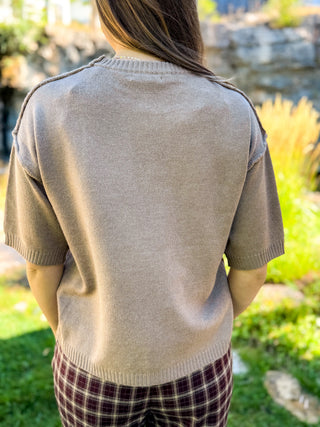 A person viewed from behind wearing a short-sleeve knit sweater in a light taupe-beige color, paired with dark plaid pants, set against a blurred outdoor garden background.