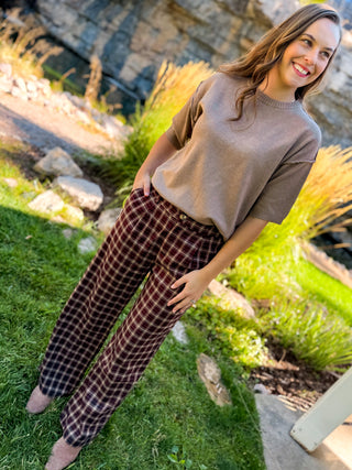 A woman standing outdoors on grass in front of a stone wall, wearing a taupe short-sleeve knit top and dark red/burgundy plaid wide-leg pants.