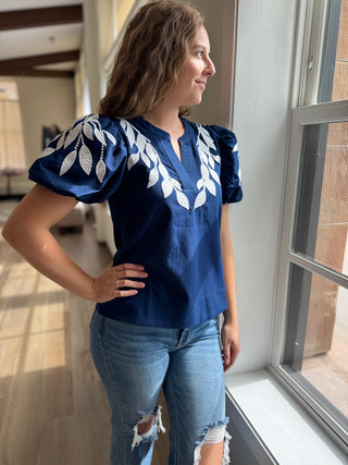 Woman wearing a blue top with white embroidery and jeans standing by a window.