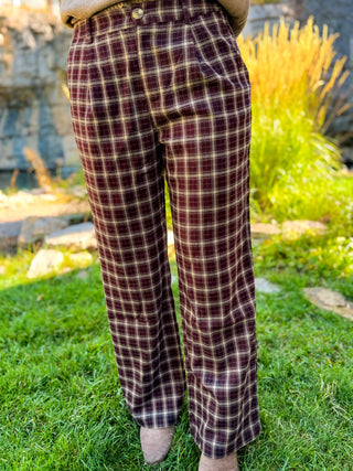 A person standing outdoors on grass wearing plaid wide-leg trousers in burgundy, beige, and dark brown, with a tan top partially visible at the waist.