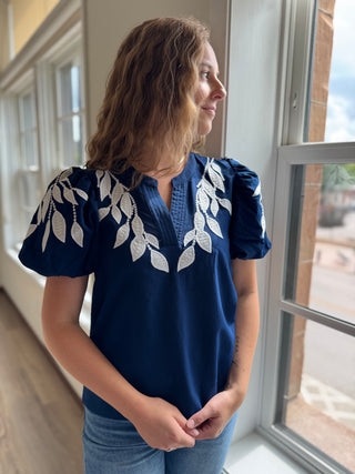 Woman wearing a navy blue blouse with white leaf patterns standing in a room with large windows.