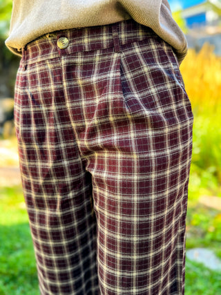 Close-up of a person wearing plaid trousers in burgundy and cream, paired with a beige sweater, outdoors with a blurred green and yellow background.
