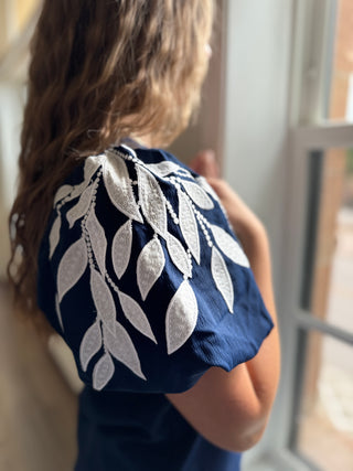 Navy blue top with white leaf patterns worn by a person, standing near a window.