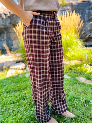 A person wearing loose, wide-leg plaid pants in a burgundy and beige pattern, standing outdoors on a grassy lawn with rocks and plants in the background.
