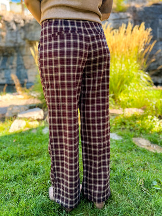 A person viewed from behind wearing wide-leg plaid pants in burgundy and beige, standing on green grass outdoors with plants and rocks in the background.