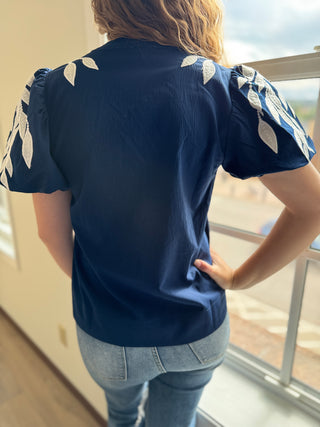 Person wearing a navy blue blouse with white leaf patterns in a room with a window.