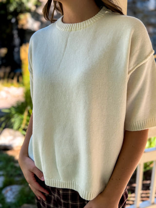 A person wearing a cream-colored short-sleeve knit sweater in an outdoor setting with blurred greenery and a white fence in the background.