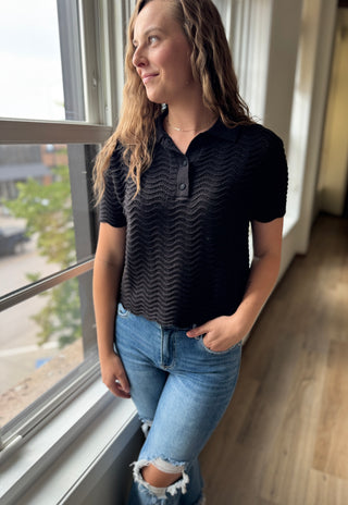 Woman wearing a black textured shirt and blue jeans standing by a window.